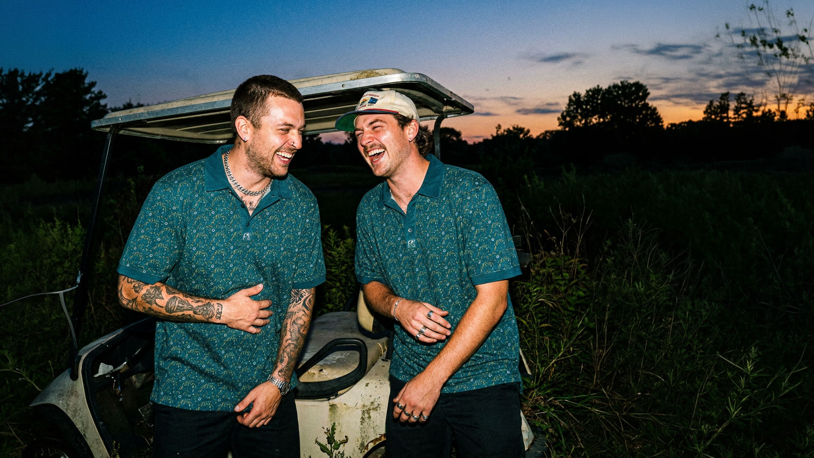 Two men in matching green shirts standing next to a vehicle in a field at dusk.
