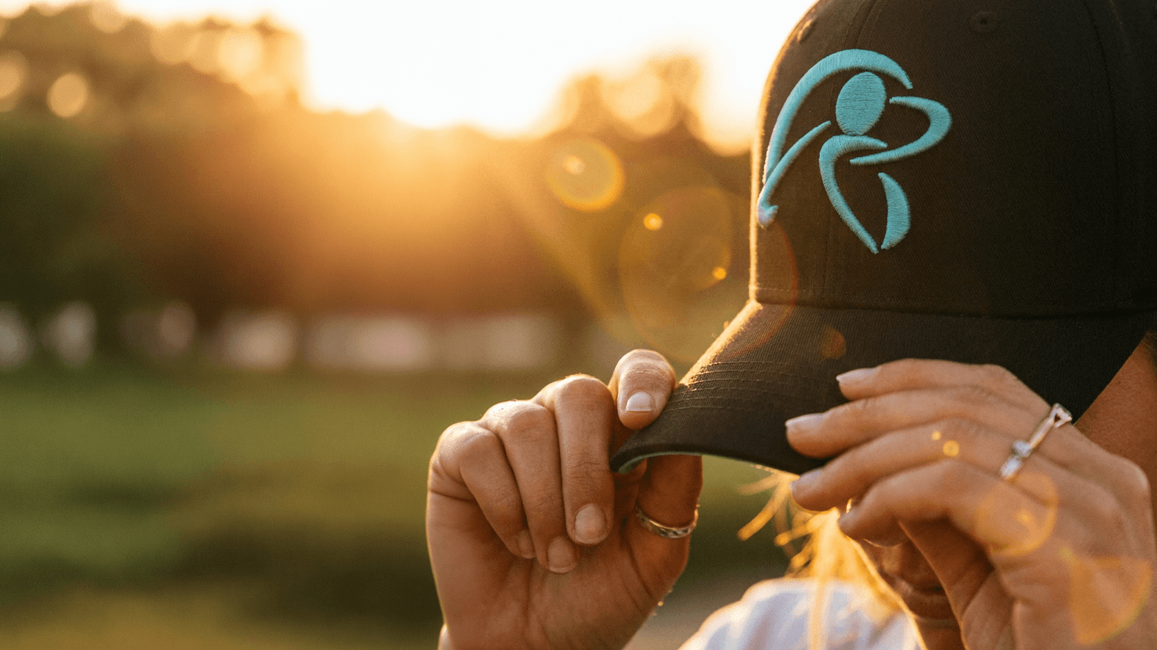 Person holding a black cap with a logo against a blurred outdoor background