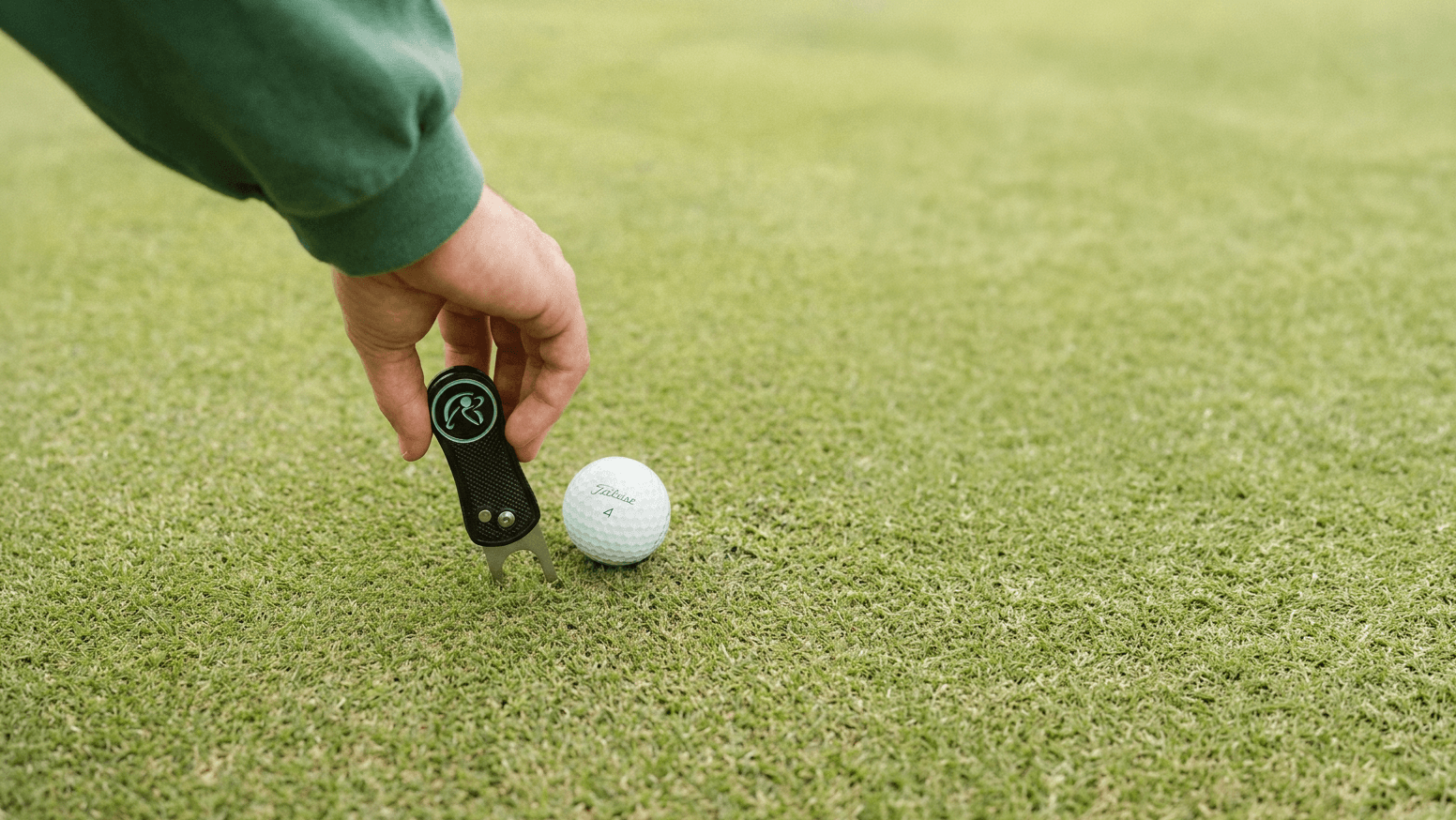 Hand holding a golf club head cover next to a golf ball on a green grass field