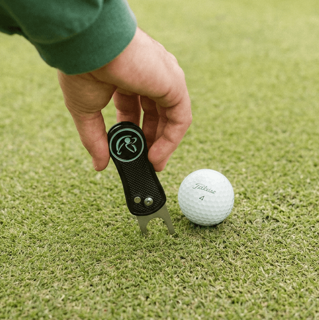 Hand holding a golf divot tool next to a golf ball on grass